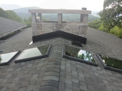 Stone chimney with new black metal flashing and four skylights on a shingle roof with a mountain view