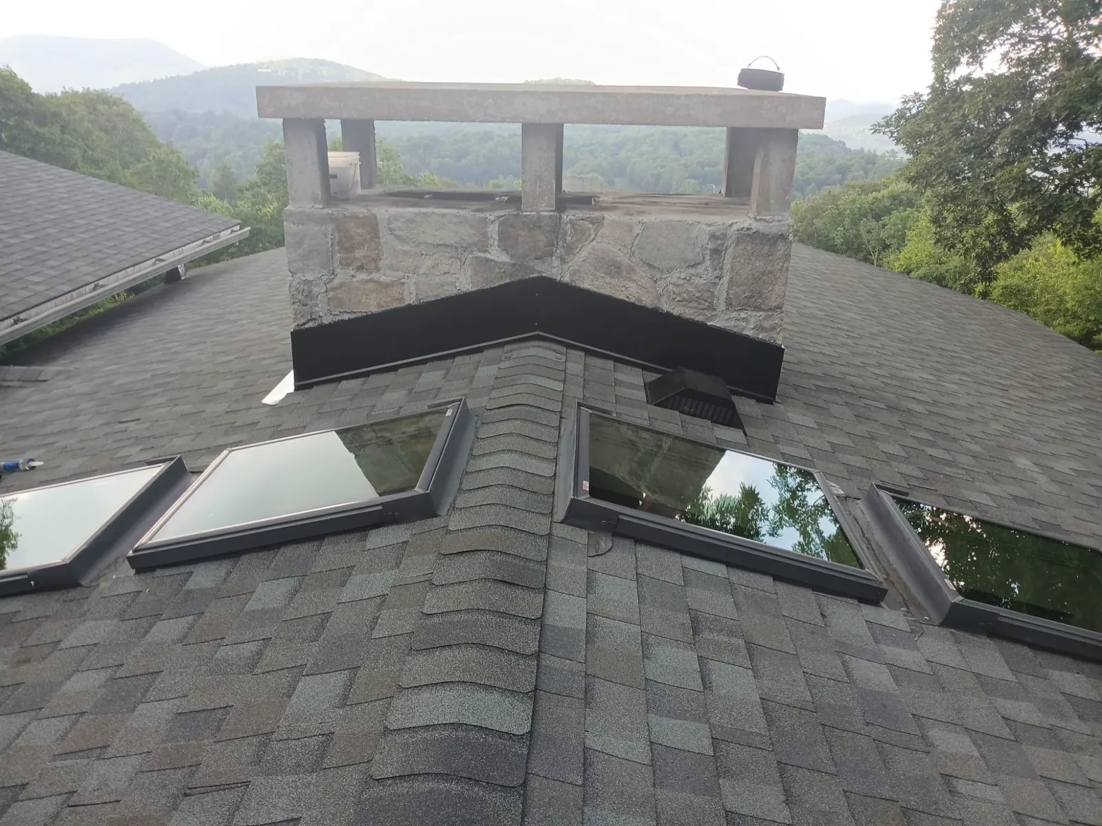 Stone chimney with new black metal flashing and four skylights on a shingle roof with a mountain view